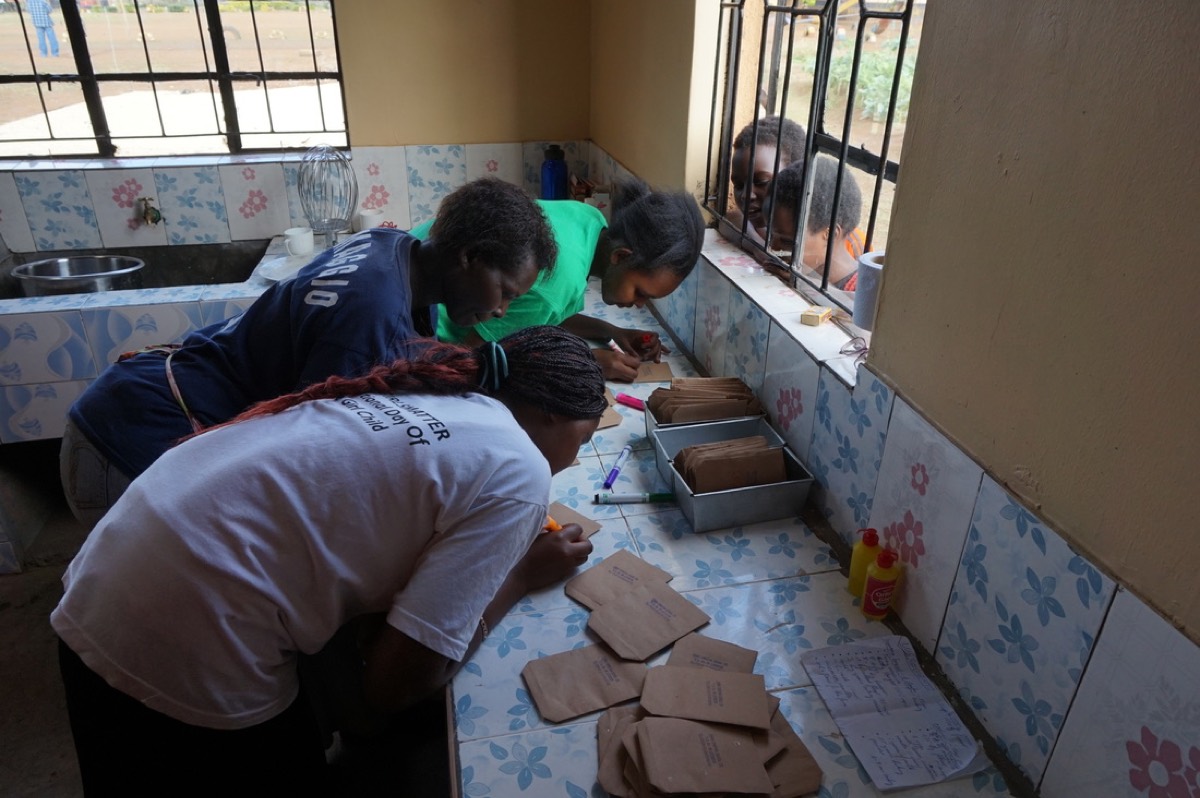 Pupils lean over a tiled counter in the school kitchen, writing labels on paper bags.